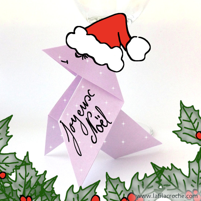 Un marque-place cocotte en origami avec un bonnet de Noël dessiné sur sa tête, des feuilles de houx en bas et les mots « Joyeux Noël » écrits sur la patte avant gauche de la cocotte