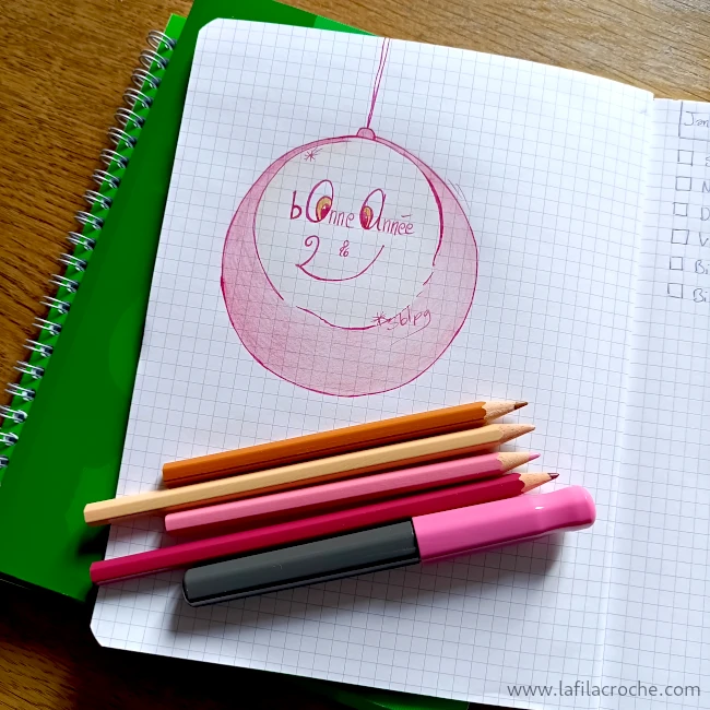 Un dessin d’une boule avec écrit bonne année 2026 dessus réalisé sur un cahier à petit carreaux. Sous le dessin on voit des crayons de couleur et un stylo-plume