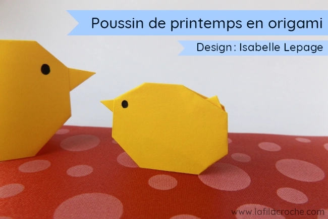 Le poussin de printemps en origami, un pliage créé par Isabelle Lepage, il est posé sur un ruban rouge à pois. À côté du poussin de printemps en papier il y a un autre poussin un peu plus grand dont on ne voit que la tête
